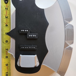 Puede incluir: Una guitarra impresa en 3D de color negro y plateado con tres selectores de pastillas negros. La guitarra tiene forma de hacha.