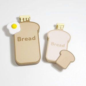 Può includere: Due contenitori a forma di pane con la scritta "Bread" stampata sopra. Un contenitore ha un uovo fritto attaccato. I contenitori sono beige e rosa chiaro, con un uovo giallo e bianco. I contenitori hanno una parte superiore gialla e ondulata.