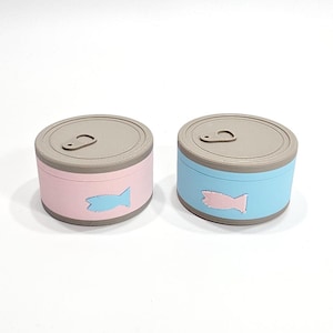 May include: Two round, colourful containers resembling cat food tins. One is pink, the other is blue, both with a grey lid and a fish-shaped emblem. The tins are designed to look like cat food.