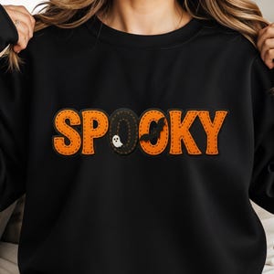 May include: Black sweatshirt featuring the word "SPOOKY" in orange, stitched lettering. The letter "O" is a black oval with a white ghost and bat design. A Halloween-themed garment.