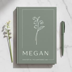 以下が含まれることがあります： 白の草花のイラストと「MEGAN」の名前が表紙に印刷されたセージグリーンのジャーナル。テキスト「CHAPTER 40: THE CONFIDENCE ERA」が下に印刷されています。お揃いのペンがジャーナルの横にあります。
