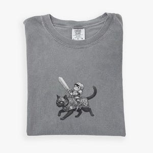 May include: Gray Comfort Colors t-shirt featuring a black and white illustration of a knight riding a black cat. The knight is holding a sword. The t-shirt is folded and has a tag in the upper right corner.