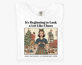 Könnte beinhalten: Weißes T-Shirt mit einer Retro-Illustration, die eine Familie beim Schmücken eines Weihnachtsbaums zeigt. Der Text "It's Beginning to Look a Lot Like Chaos" und "And Patience is Running Low" sind oberhalb und unterhalb der Illustration gedruckt.