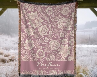 Può includere: Coperta malva con un motivo floreale color crema. La coperta ha un bordo con frange e la parola "Mother" è stampata su una fascia in basso, insieme ai nomi. La coperta è appesa all'aperto.