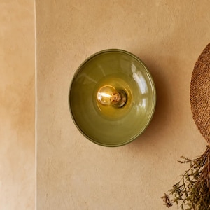 Può includere: Una lampada da parete circolare con finitura verde oliva lucida e una lampadina a luce calda esposta al centro. L'apparecchio è montato su una parete beige strutturata, accanto a un cappello di paglia e un mazzetto di erbe essiccate.