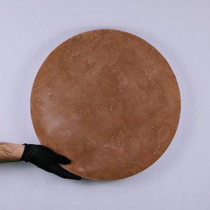 Pode incluir: Uma grande tábua de servir redonda de pedra castanho-avermelhada. A tábua tem uma superfície texturizada e é segurada por uma mão enluvada contra um fundo branco. A tábua tem cerca de 46 cm de diâmetro.