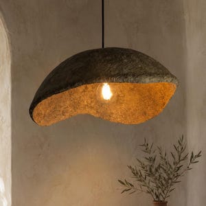 May include: A pendant light fixture featuring a large, irregularly shaped, dark gray textured shade with a warm, golden-yellow interior finish. The lamp hangs from a thin black cord and contains a single glowing light bulb in the center.