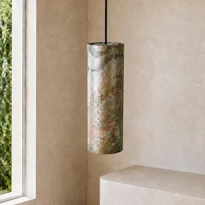 May include: A cylindrical pendant light fixture featuring a textured, mottled surface with variegated shades of green, brown, and metallic copper tones. The fixture is suspended from a thin, straight black cord attached to the top center.