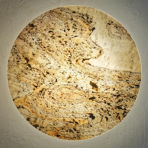 Puede incluir: Una obra de arte mural redonda y decorativa con apariencia de piedra natural. La superficie presenta una mezcla de tonos beige, marrón y tostado, creando un efecto marmolado. La obra está iluminada con una luz suave y difusa en el borde.