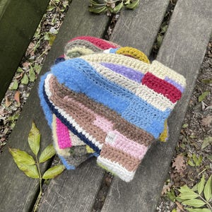 Könnte beinhalten: Ein mehrfarbiger Häkelhut mit Patchwork-Design. Der Hut hat blaue, rosa, braune und weiße Streifen. Der Hut liegt auf einer Holzbank. Der Hut hat die Buchstaben 'MAF'.