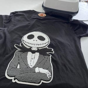 May include: A black t-shirt featuring a large, textured chenille patch of a skeleton character in a suit, with a smaller football-shaped patch near the neckline. A white and pink heat press machine sits at the top of the shirt on the table.