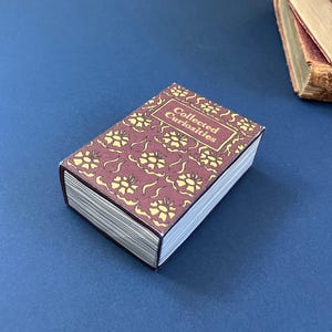 Puede incluir: Una pequeña caja rectangular con el título "Collected Curiosities" en la portada. La portada es de color burdeos con un patrón floral amarillo repetido. La caja está sobre una superficie azul, con otros libros visibles en el fondo. Mide unos 10 cm de largo.