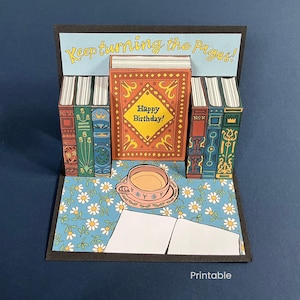 May include: A pop-up birthday card featuring a central book with a "Happy Birthday!" message, flanked by stacks of colorful books. A teacup and saucer sit on a floral patterned surface, with the text "Keep turning the pages!" above.
