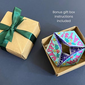 May include: A square kraft paper gift box with a dark green satin ribbon bow, alongside an open kraft box containing a folded paper ornament with the text "HAPPY HOLIDAYS!" in pink, decorated with holly leaves, berries, and yellow stars.