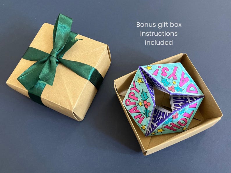 May include: A square kraft paper gift box with a dark green satin ribbon bow, alongside an open kraft box containing a folded paper ornament with the text "HAPPY HOLIDAYS!" in pink, decorated with holly leaves, berries, and yellow stars.