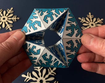 Puede incluir: Un adorno geométrico de papel con forma hexagonal y una abertura central. La superficie está decorada con un patrón repetitivo de motivos de copos de nieve y estrellas blancas sobre un fondo azul cerceta vibrante en 3D.