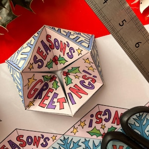 Könnte beinhalten: Ein gefalteter Papier-Wahrsager mit den Worten "SEASON'S GREETINGS!" in Rot und Lila. Das Design umfasst Stechpalmen, Sterne sowie blaue und gelbe Akzente. Ein Metalllineal und eine Schere sind ebenfalls sichtbar.