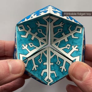 Könnte beinhalten: Ein blau-weißes geometrisches Zappelspielzeug mit Schneeflockenmustern. Das Spielzeug besteht aus gefaltetem Papier und wird von zwei Händen gehalten. Der Text "Printable fidget toy" ist auf dem Bild sichtbar.