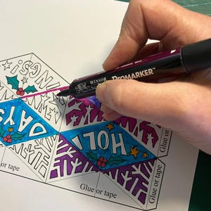 May include: A dark purple Promarker felt-tip pen coloring a paper template featuring diamond-shaped sections with the text HOLIDAYS, holly leaves, berries, and star patterns. The paper includes printed instructions labeled Glue or tape.