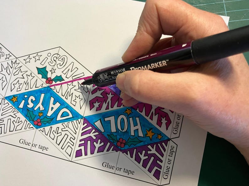 May include: A dark purple Promarker felt-tip pen coloring a paper template featuring diamond-shaped sections with the text HOLIDAYS, holly leaves, berries, and star patterns. The paper includes printed instructions labeled Glue or tape.