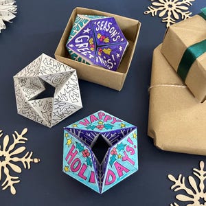 May include: Three geometric paper ornaments with triangular facets. One is black and white with star patterns, one is colored blue and purple with the text "SEASON'S GREETINGS", and one is blue and pink with the text "HAPPY HOLIDAYS".
