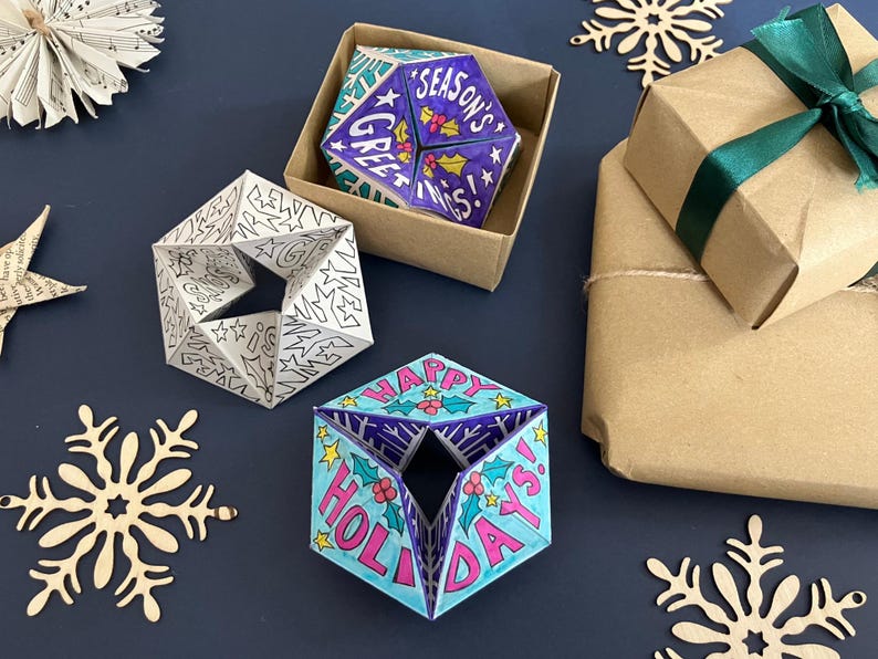 May include: Three geometric paper ornaments with triangular facets. One is black and white with star patterns, one is colored blue and purple with the text "SEASON'S GREETINGS", and one is blue and pink with the text "HAPPY HOLIDAYS".