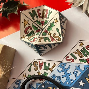 Könnte beinhalten: Handgefertigte Weihnachtsdekoration aus Papier mit dem Aufdruck „MERRY CHRISTMAS!“ auf den Seiten. Die Dekoration zeigt Stechpalmen- und Sternillustrationen. Weitere Artikel sind Geschenkpapier, Schere und eine Geschenkbox.