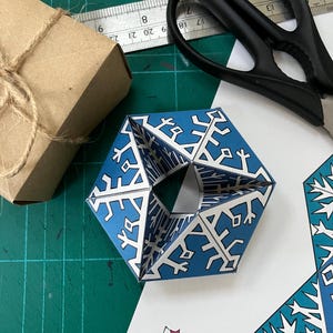 Könnte beinhalten: Eine blaue und weiße geometrische Papierskulptur mit Schneeflockenmuster. Ein braunes Papierpaket, das mit Bindfaden verschnürt ist, eine Schere und ein Lineal sind ebenfalls sichtbar. Die Skulptur befindet sich auf einer grünen Schneidematte.
