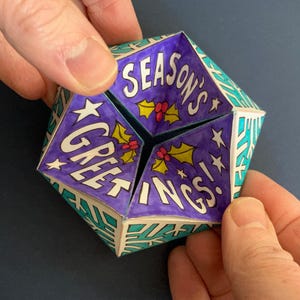 May include: A folded paper fortune teller toy featuring a purple interior with the text "SEASON'S GREETINGS!" in white letters, accented by green holly leaves and red berries. The exterior displays a geometric pattern of white snowflakes on a teal background.