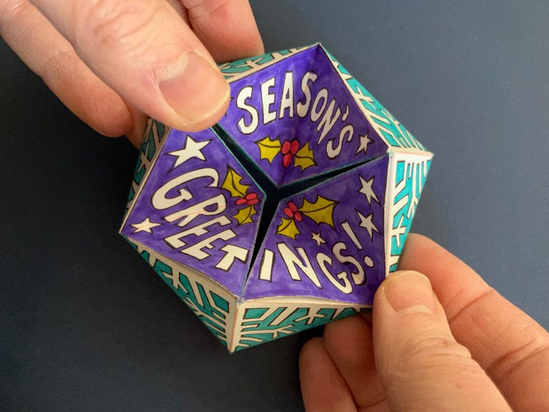 May include: A folded paper fortune teller toy featuring a purple interior with the text "SEASON'S GREETINGS!" in white letters, accented by green holly leaves and red berries. The exterior displays a geometric pattern of white snowflakes on a teal background.