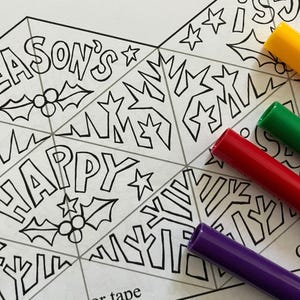 May include: A black and white line art coloring sheet featuring triangular segments with the text SEASON'S and HAPPY, decorated with holly berries, leaves, and stars. Several colorful felt-tip markers in yellow, green, red, blue, and purple are placed nearby.