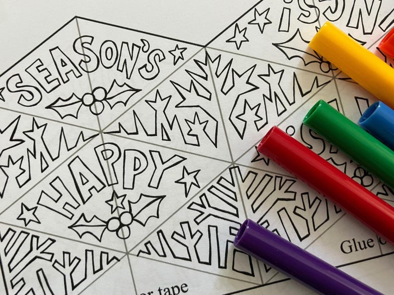May include: A black and white line art coloring sheet featuring triangular segments with the text SEASON'S and HAPPY, decorated with holly berries, leaves, and stars. Several colorful felt-tip markers in yellow, green, red, blue, and purple are placed nearby.