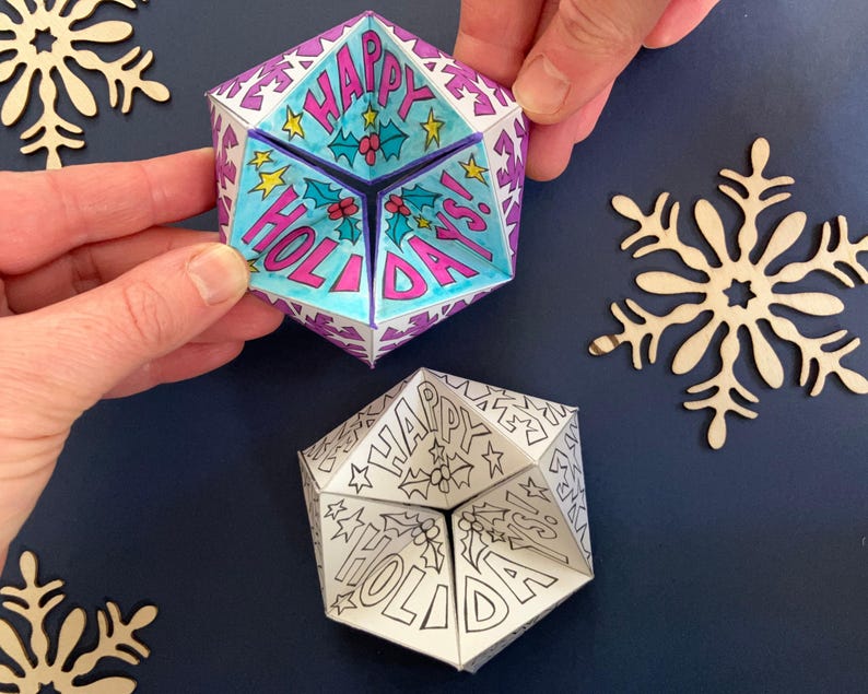 Two printable paper kaleidocycles. The visible face of each shows hand drawn decorations and the words 'Happy Holidays!' One has been printed in black and white ready to be coloured-in and the other has been coloured so that the background is blue.