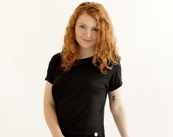 May include: A woman with long, curly red hair is wearing a black, short-sleeved t-shirt with a side slit. She is wearing black pants and has a small tattoo on her arm.