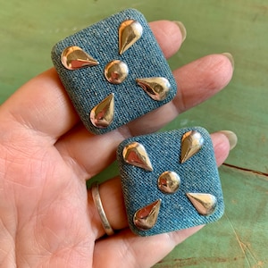 May include: A pair of denim earrings with gold metal accents. The earrings are square shaped and have a denim fabric texture. The gold accents are in the shape of teardrops and are arranged in a starburst pattern.