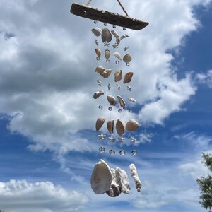 May include: A hanging mobile featuring a horizontal piece of weathered driftwood suspended by a jute rope. Four vertical strands hang from the wood, decorated with various white seashells, small white starfish, and clear glass beads throughout.