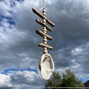 Könnte beinhalten: Ein hängendes Dekorationsobjekt mit vier horizontal angeordneten hellbraunen Treibholzstücken, die durch kleine runde Holzperlen auf einer Juteschnur getrennt sind und unten mit einer großen weißen Muschel vertikal abgeschlossen werden.