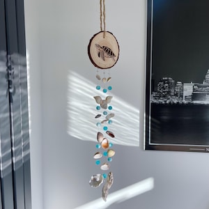 May include: A hanging wooden slice with a laser-etched turtle design, strung with seashells, turquoise beads, and natural twine. This coastal-themed mobile is a decorative accent for any room.