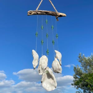 May include: A hanging mobile featuring a piece of driftwood at the top with a jute rope hanger. Five clear nylon strings hang from the wood, each decorated with small, faceted green glass beads and finished with a white oyster shell at the bottom.