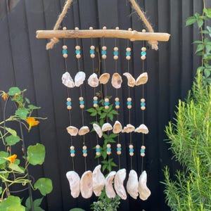 May include: A hanging wall decoration featuring a horizontal wooden branch suspended by a jute rope. Nine vertical strings hang from the branch, each adorned with alternating light blue and natural wooden beads and white seashells.