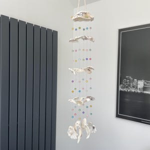 May include: A decorative hanging mobile composed of several off-white ceramic shell shapes. The shells are strung together with clear thread, interspersed with colourful beads. The mobile is suspended from a natural rope.