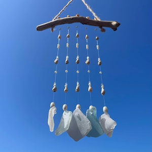 Peut inclure: Un carillon fait à la main avec une branche de bois flotté, orné de cordes de petits coquillages, de perles et de morceaux de verre de mer blancs, bleus et transparents. Le carillon est suspendu devant un ciel bleu clair.