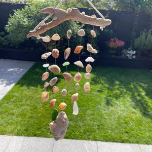 Op de afbeelding: Een hangende mobiel gemaakt van een stuk drijfhout dat aan een touw hangt. Aan het hout hangen meerdere dunne koorden, elk versierd met diverse natuurlijke schelpen in tinten beige, wit en bruin, afgewisseld met kleine stukjes groen en wit zeeglas.