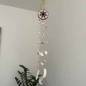 Puede incluir: Móvil colgante con diseño de sol y conchas marinas. La parte superior es un disco de madera con un motivo de sol. Cadenas de conchas marinas y cuentas doradas cuelgan debajo. El móvil está suspendido por un cordón dorado.