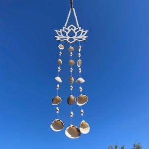May include: A white lotus flower wind chime with hanging strands of seashells and small beads. The wind chime is suspended against a clear blue sky, creating a coastal, beachy aesthetic.