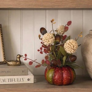 May include: A decorative pumpkin-shaped vessel in shades of red and green, topped with an arrangement of dried cream-colored spherical flowers, brown acorns, and small green leaves, displayed as a centerpiece on a wooden shelf.