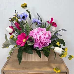 May include: A vibrant floral arrangement featuring a large pink peony as the focal point, surrounded by an assortment of colorful blooms. The flowers are arranged in a light-colored rectangular wooden container, placed on a wooden cabinet.