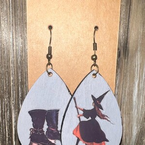 May include: Teardrop-shaped earrings with a witch theme. One earring features a pair of black boots, the other a witch on a broom. The earrings have a light gray background and bronze-colored ear wires.