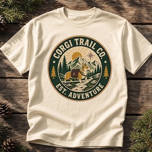Può includere: Una t-shirt color crema con una grafica circolare dal bordo verde. Il disegno raffigura un Corgi con uno zaino su un sentiero, con montagne, pini, un sole e una stella polare. Il testo riporta CORGI TRAIL CO. EST. ADVENTURE.