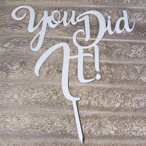 May include: A white cake topper with the phrase "You Did It!" in a flowing script. The topper is made from a solid material and features a pointed stake at the base for securing it into a cake or similar treat.